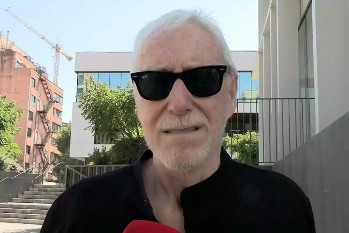 Josep María Mainat més gran amb barba i ulleres de sol negres dret a l’aire lliure davant d’un tanatori