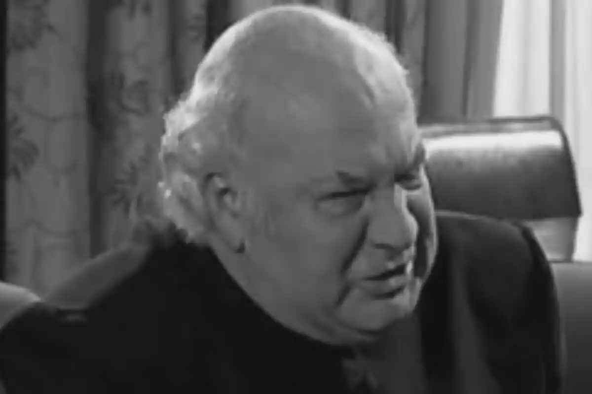 Juan Mandli, home gran i calb amb expressió seriosa assegut en un sofà vermell dins d’una habitació amb cortines clares a La que se avecina en una imatge en blanc i negre