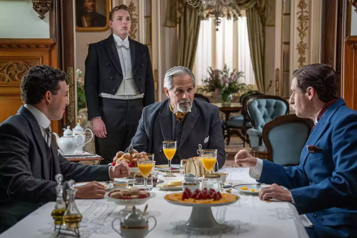 Tres hombres elegantemente vestidos conversan en una mesa de desayuno con jugo de naranja y pasteles mientras un mayordomo de pie los observa en una lujosa sala decorada con muebles clásicos. Captura de 'La Promesa' Tres hombres elegantemente vestidos conversan en una mesa de desayuno con jugo de naranja y pasteles mientras un mayordomo de pie los observa en una lujosa sala decorada con muebles clásicos. Captura de 'La Promesa'