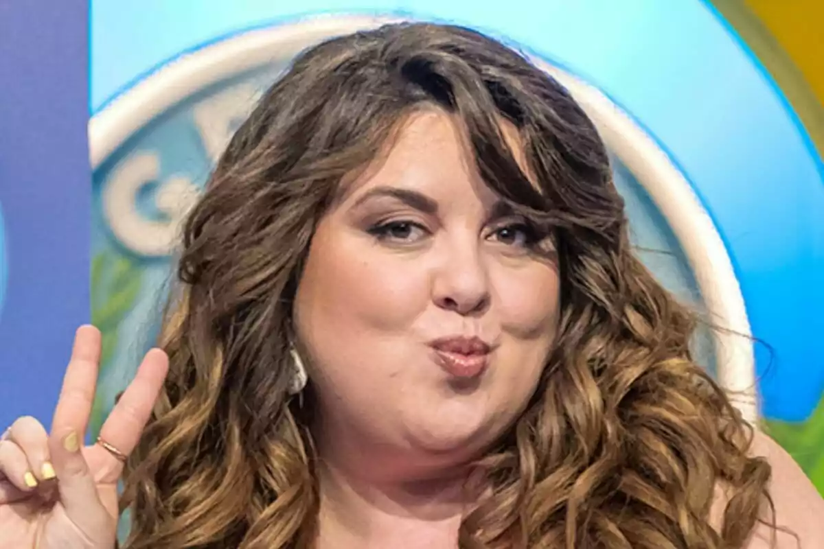 Lalachus de cabello largo y ondulado haciendo un gesto de beso y señal de paz con los dedos frente a un fondo colorido en El Grand Prix.