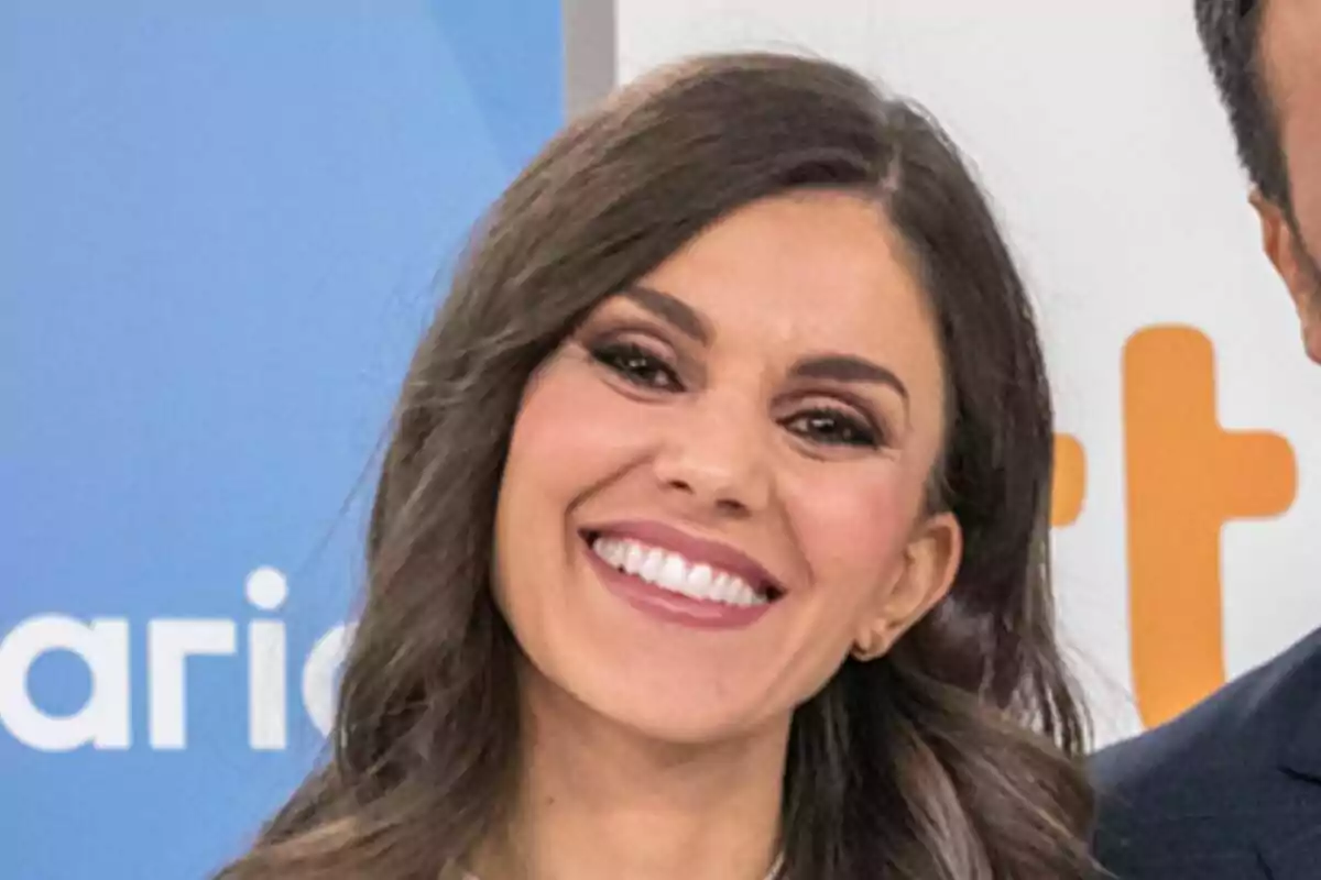 Lorena Baeza de cabello castaño largo y ondulado sonriendo frente a un fondo azul y blanco del Telediario con letras naranjas y blancas. Lorena Baeza de cabello castaño largo y ondulado sonriendo frente a un fondo azul y blanco del Telediario con letras naranjas y blancas.