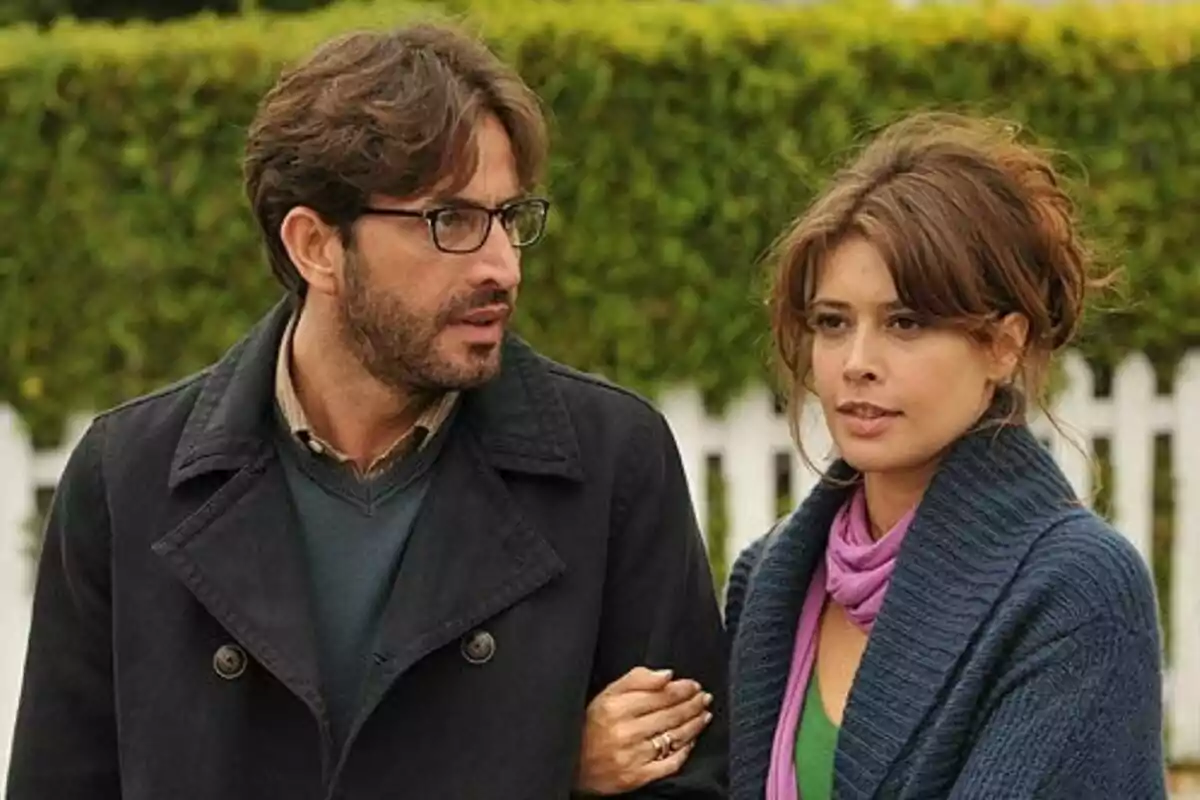Angie Cepeda y Antonio Garrido caminando al aire libre con expresión seria y fondo de arbustos y una cerca blanca en Los Protegidos.