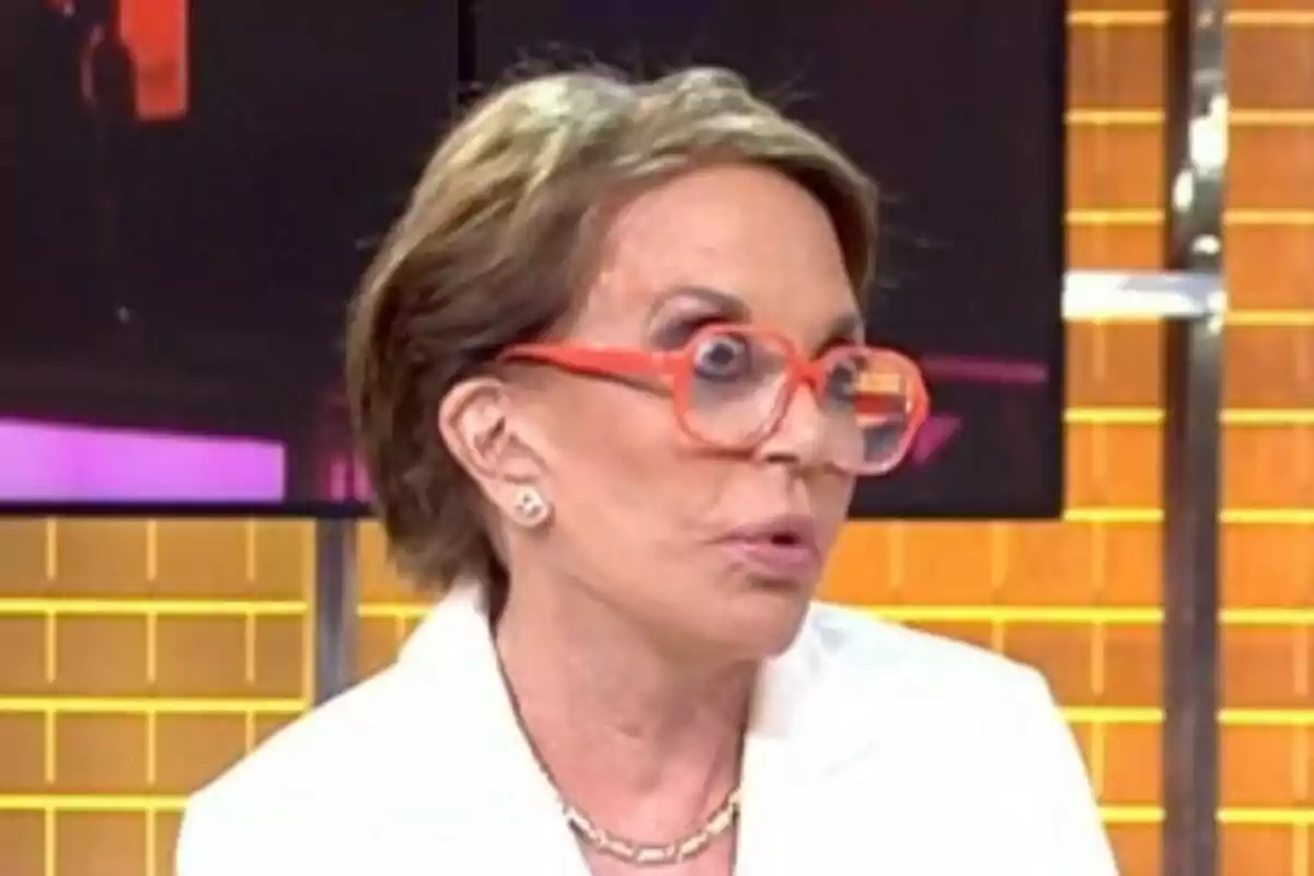 María Eugenia Yagüe de cabello corto con gafas grandes naranjas y expresión de sorpresa en un set de Fiesta con fondo de colores cálidos