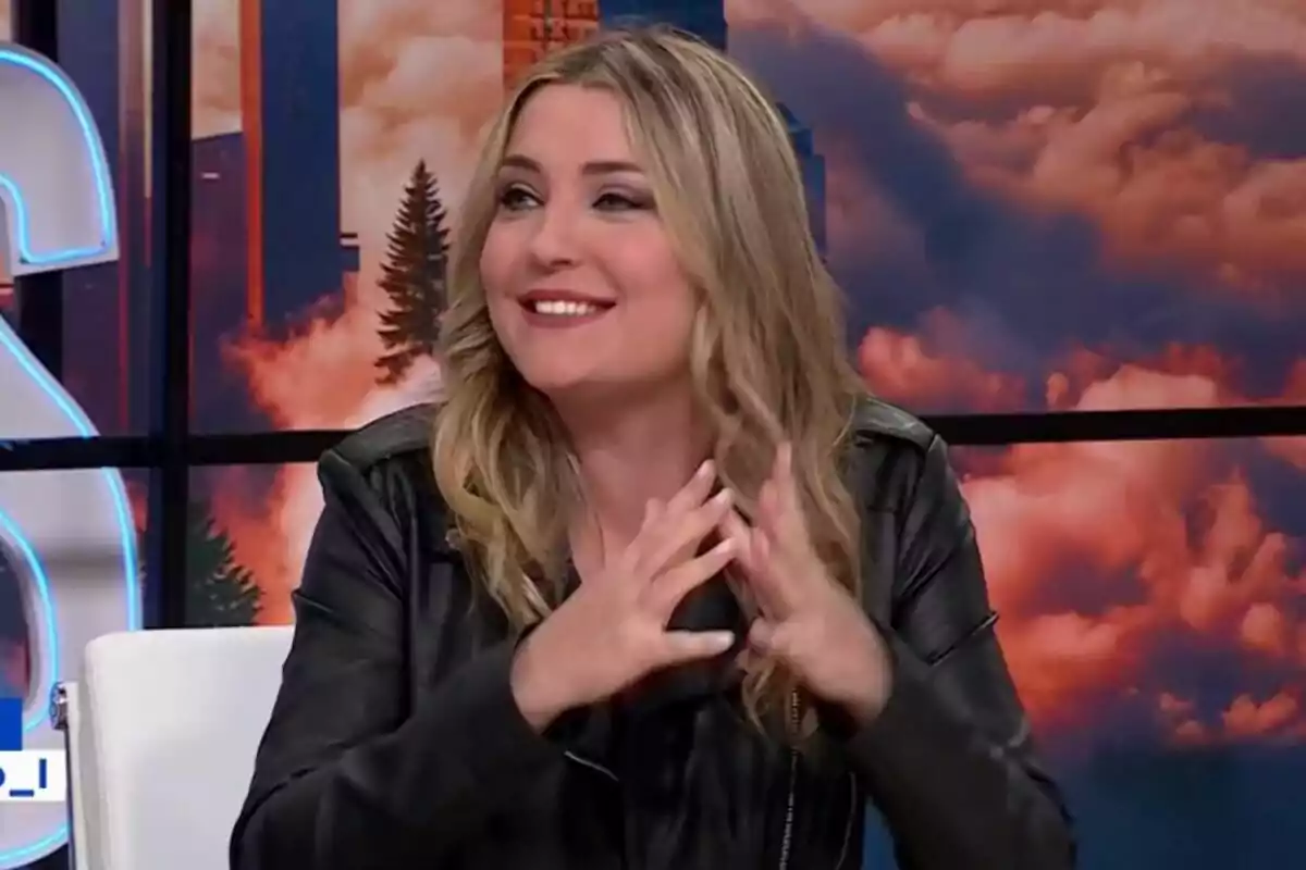 Marina Lobo rubia sonriente con chaqueta de cuero negra sentada en un set de Malas Lenguas con fondo de nubes y edificios Marina Lobo rubia sonriente con chaqueta de cuero negra sentada en un set de Malas Lenguas con fondo de nubes y edificios