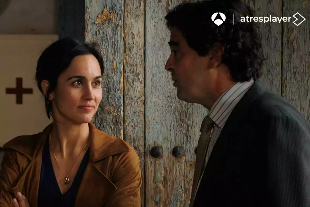 Megan Montaner y un hombre conversan frente a una puerta de madera desgastada, ella sonríe levemente mientras lo escucha atentamente en Entre Tierras.