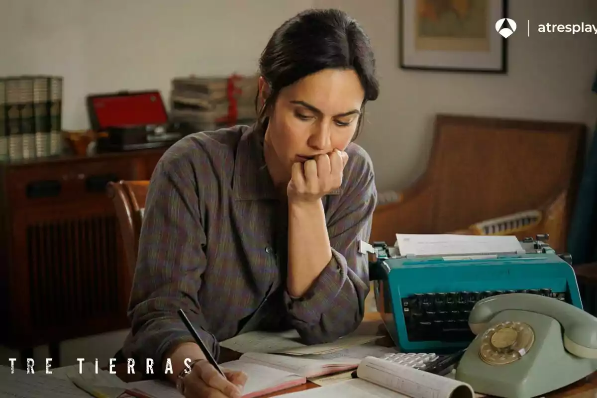 Megan Montaner pensativa sentada en un escritorio con una máquina de escribir azul, un teléfono antiguo y varios papeles y libros en Entre Tierras Megan Montaner pensativa sentada en un escritorio con una máquina de escribir azul, un teléfono antiguo y varios papeles y libros en Entre Tierras