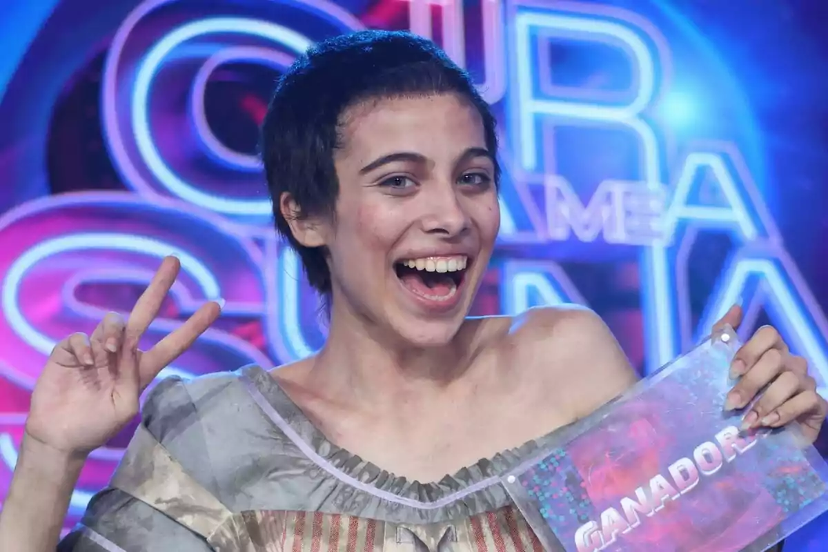 Melani sonriente con el cabello corto haciendo la señal de la paz y sosteniendo un cartel que dice ganador en un escenario colorido de Tu Cara Me Suena.