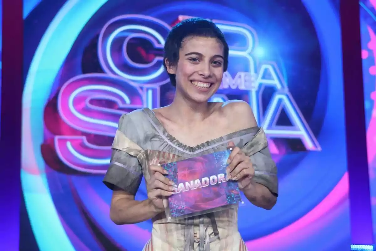 Melani sonriente sostiene un cartel con la palabra ganadora en un set de televisión colorido de Tu Cara Me Suena Melani sonriente sostiene un cartel con la palabra ganadora en un set de televisión colorido de Tu Cara Me Suena
