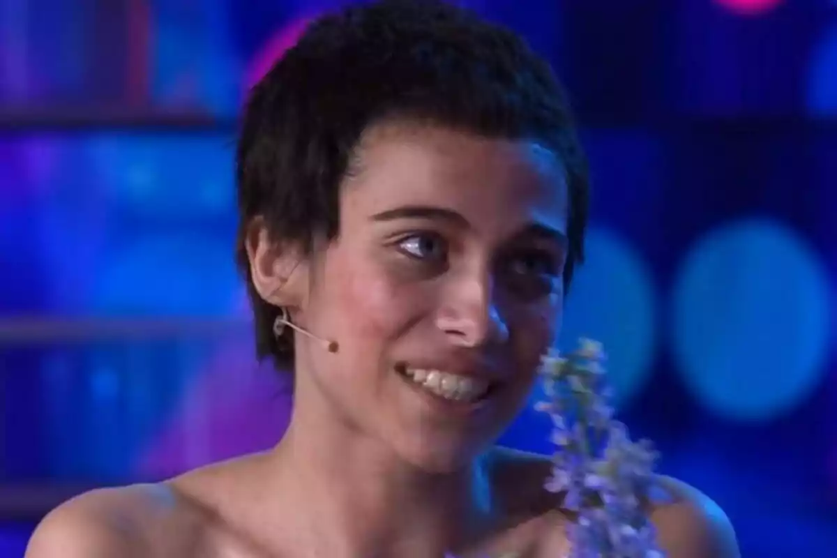 Melani de cabell curt i fosc somrient en un escenari il·luminat de Tu Cara Me Suena amb llums blaves i violetes, portant un micròfon de diadema i sostenint flors.