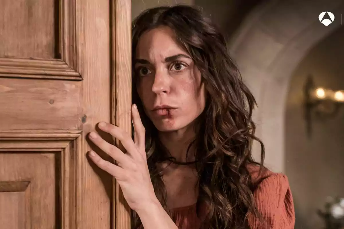 Mujer con expresión preocupada y herida en el labio se asoma detrás de una puerta de madera en Las hijas de la criada Mujer con expresión preocupada y herida en el labio se asoma detrás de una puerta de madera en Las hijas de la criada