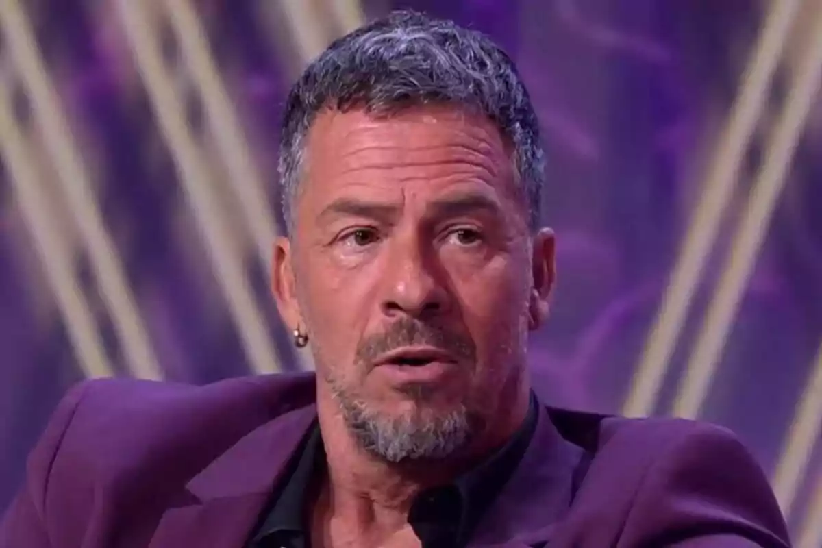 Nacho Palau de cabello corto y barba con saco morado sentado en un set de De Viernes con fondo púrpura