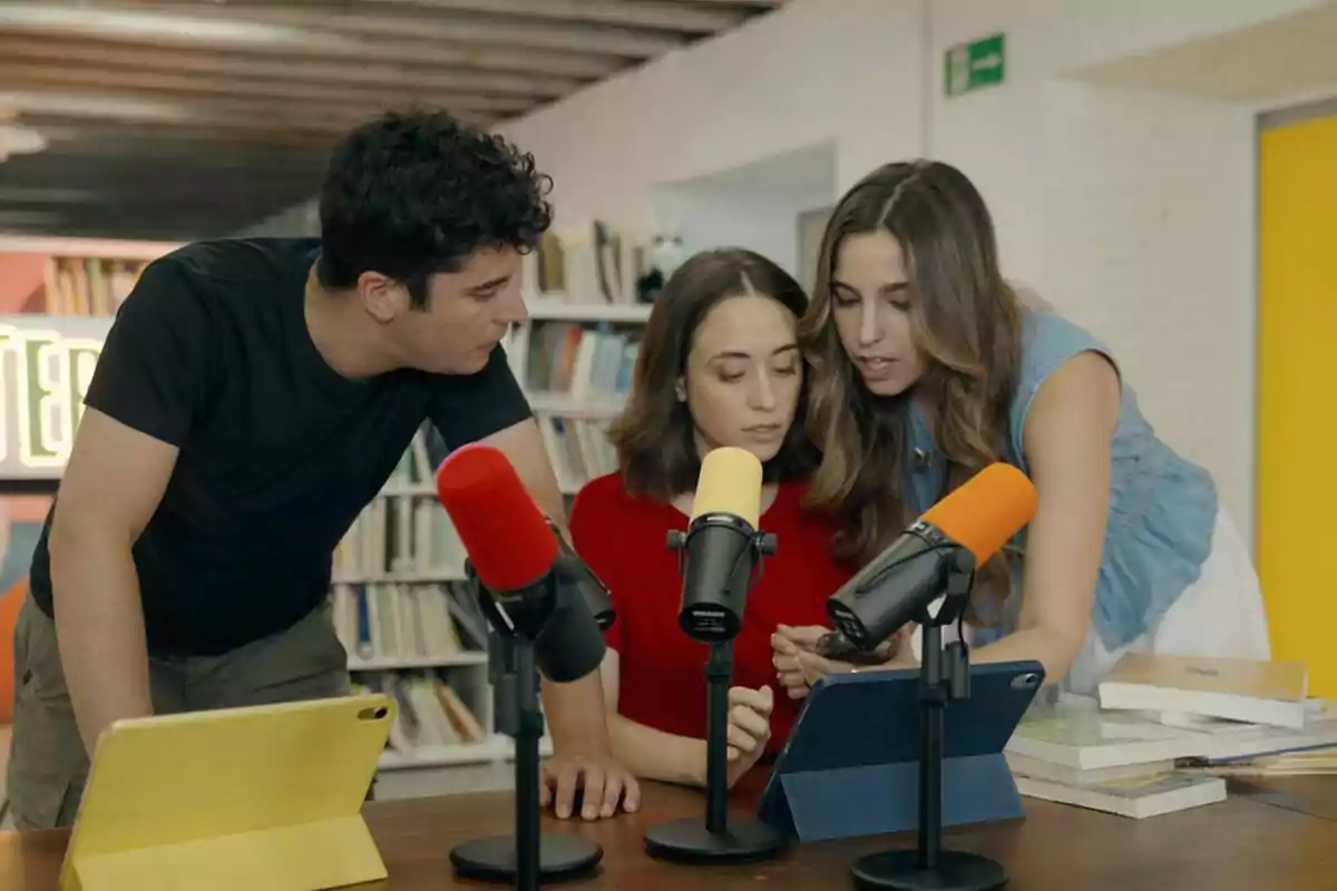 Emilio Doménech juntament amb Albanta San Román i Alba Leiva revisen una tauleta en una taula amb micròfons de colors i llibres en un ambient de biblioteca Emilio Doménech juntament amb Albanta San Román i Alba Leiva revisen una tauleta en una taula amb micròfons de colors i llibres en un ambient de biblioteca