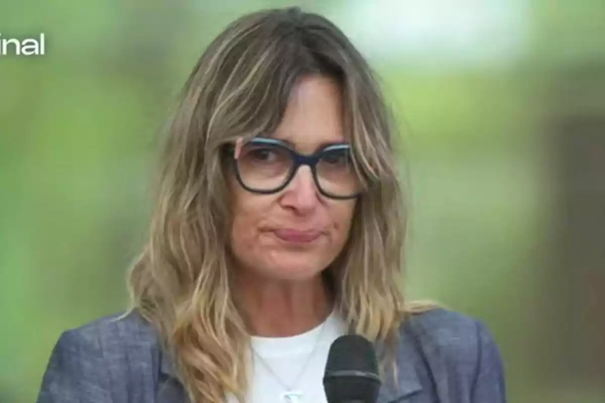 Noemí Galera de cabello rubio y gafas sostiene un micrófono con expresión seria y fondo desenfocado en Operación Triunfo.