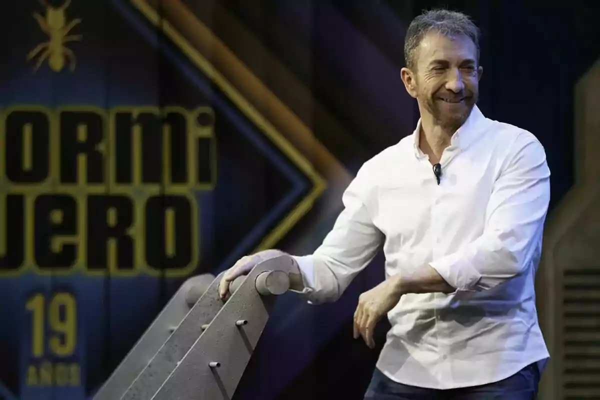 Pablo Motos de camisa blanca sonriendo en un set de El Hormiguero con el logo de un programa al fondo Pablo Motos de camisa blanca sonriendo en un set de El Hormiguero con el logo de un programa al fondo