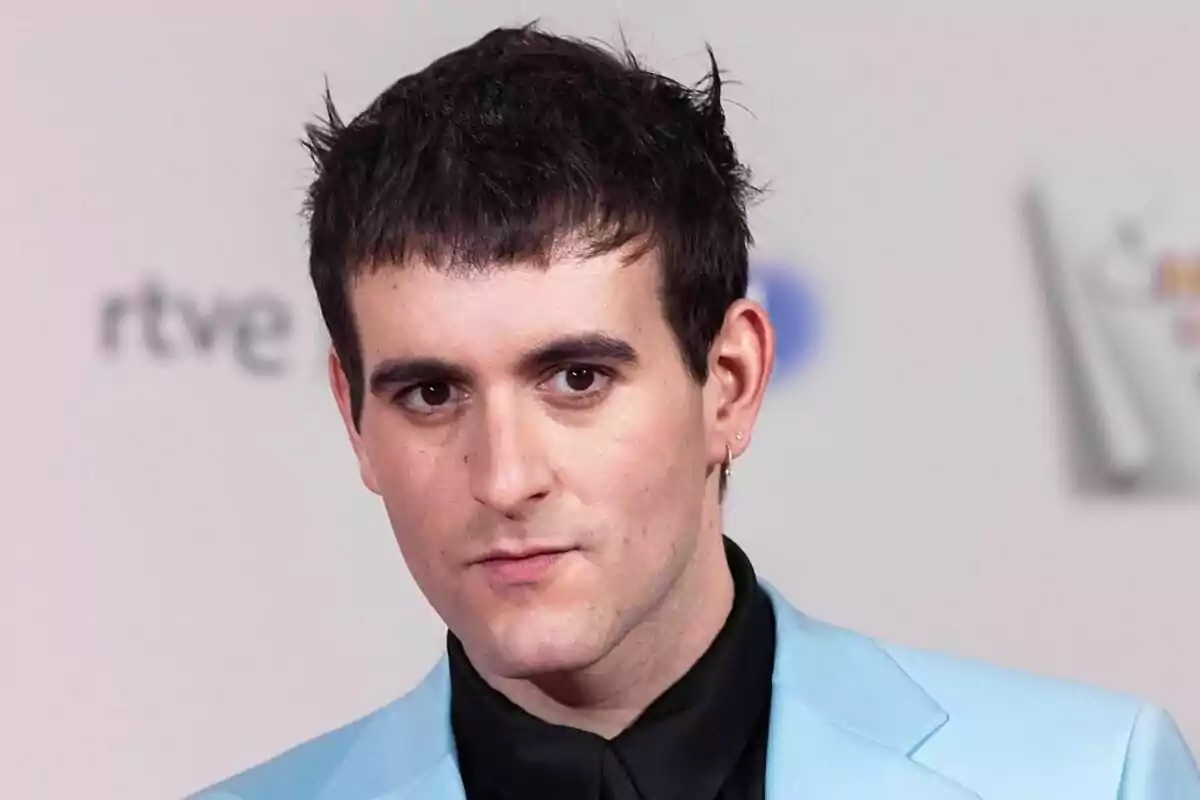 Palomo Spain de cabello corto y oscuro con chaqueta azul claro y camisa negra posando frente a un fondo claro con logotipos desenfocados en Maestros de la Costura.