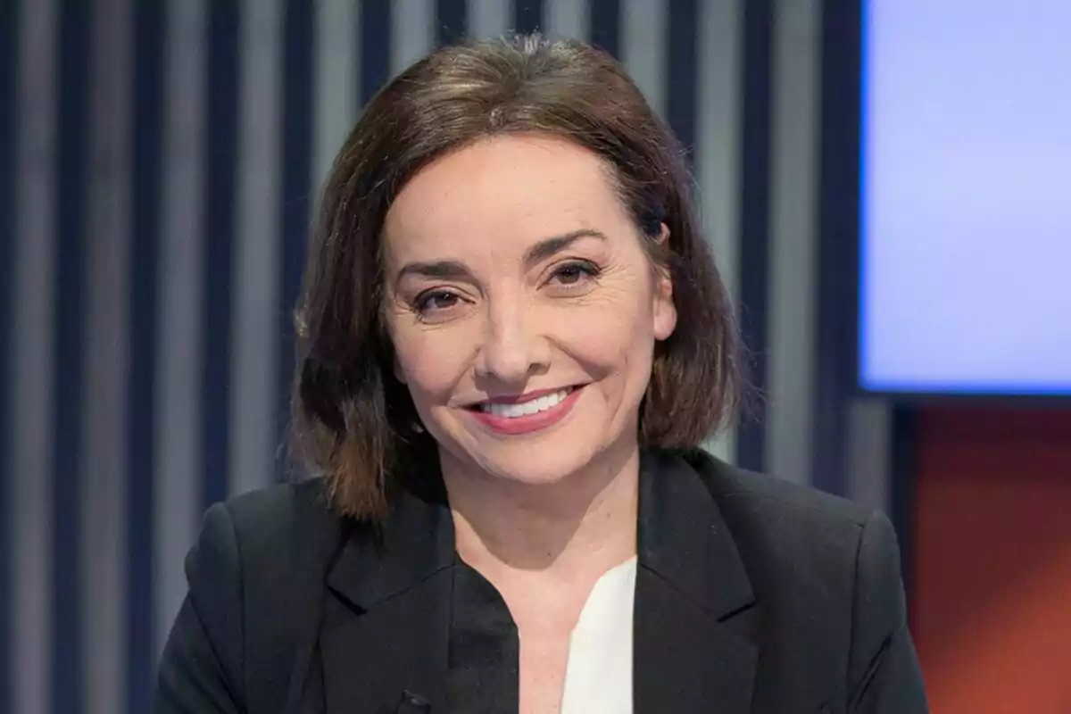 Pepa Bueno de cabello corto y oscuro con saco negro y blusa blanca sonriendo frente a un fondo con líneas verticales.