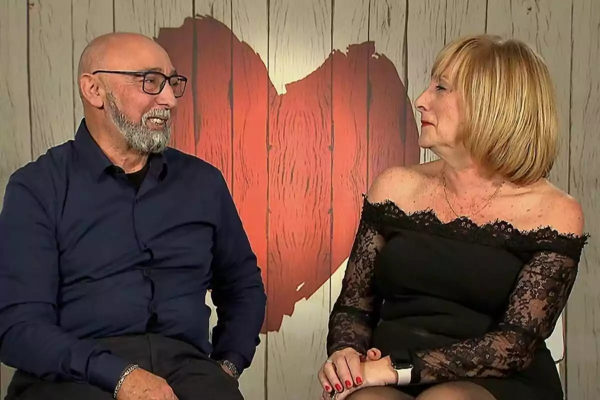 Un home i una dona grans, en Lluís i la Pilar, asseguts cara a cara somrient, amb un fons de fusta clara i un gran cor vermell darrere d’ells a First Dates.