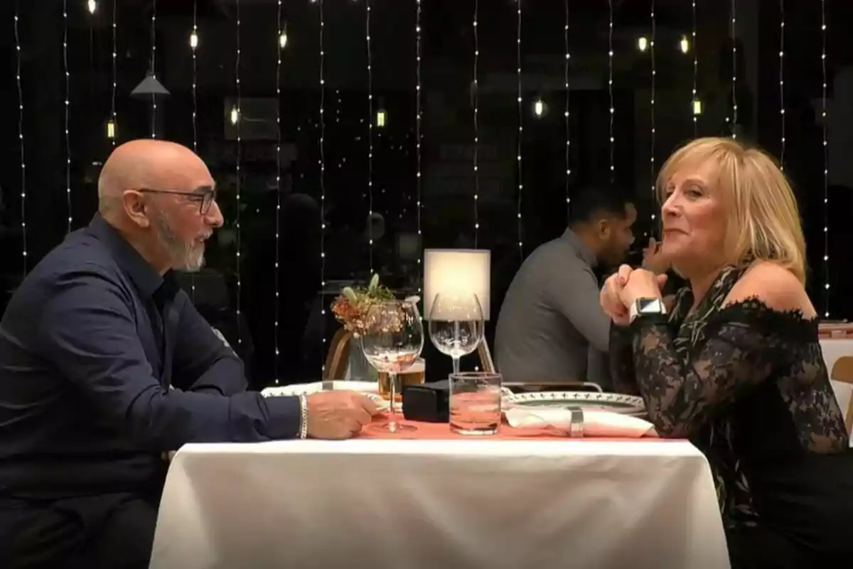 Una parella de persones grans, la Pilar i en Lluís, assegudes cara a cara en una taula de restaurant de First Dates elegantment decorada, conversant durant una cita nocturna amb llums de fons. Una parella de persones grans, la Pilar i en Lluís, assegudes cara a cara en una taula de restaurant de First Dates elegantment decorada, conversant durant una cita nocturna amb llums de fons.