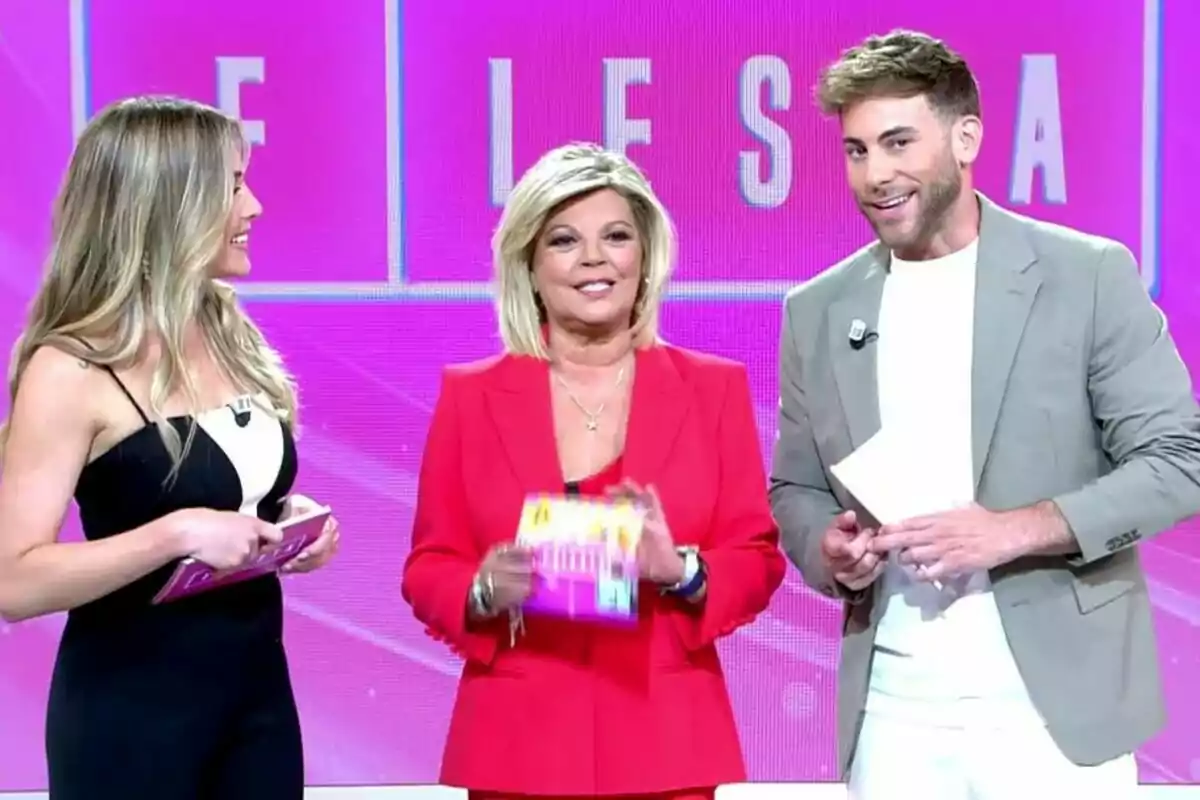 Tres presentadors de televisió, Álex Blanquer, Terelu Campos i César Muñoz, somriuen en un plató de Fiesta amb un fons rosa mentre sostenen targetes a les mans.