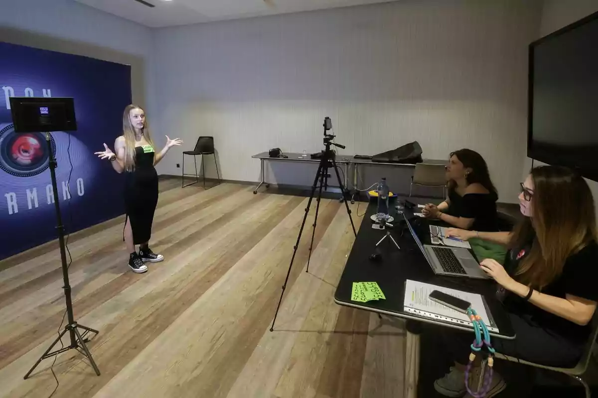 Una joven participa en un casting de Gran Hermano frente a Teresa Colomina y otra mujer que la evalúan mientras toman notas en una computadora y la graban con una cámara en una sala con piso de madera Una joven participa en un casting de Gran Hermano frente a Teresa Colomina y otra mujer que la evalúan mientras toman notas en una computadora y la graban con una cámara en una sala con piso de madera