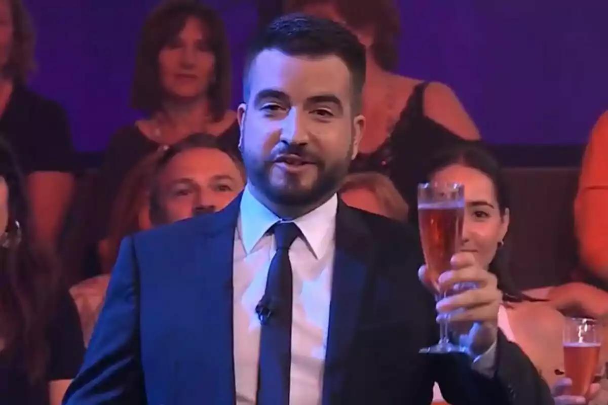 Ricard Ustrell amb barba i vestit sostenint una copa de vi escumós en un estudi de televisió amb públic al fons a Col·lapse Ricard Ustrell amb barba i vestit sostenint una copa de vi escumós en un estudi de televisió amb públic al fons a Col·lapse