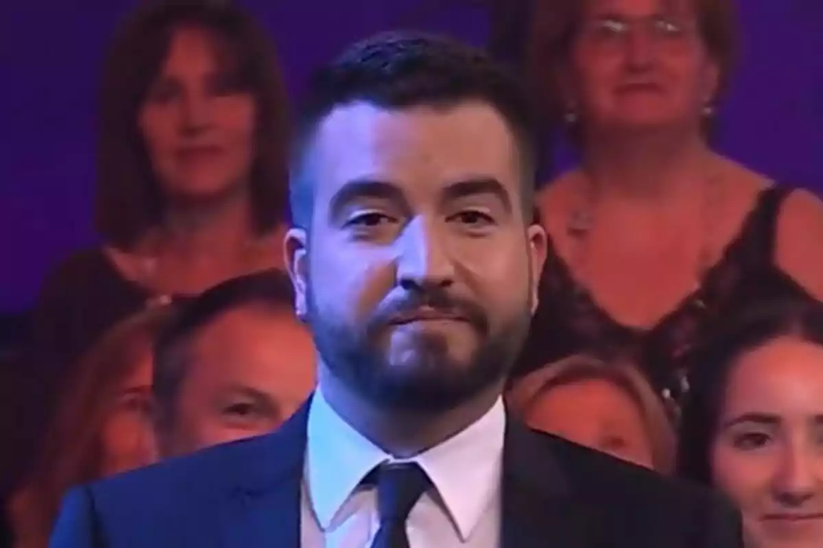 Ricard Ustrell amb barba i vestit fosc en primer pla amb públic desenfocat al fons a Col·lapse