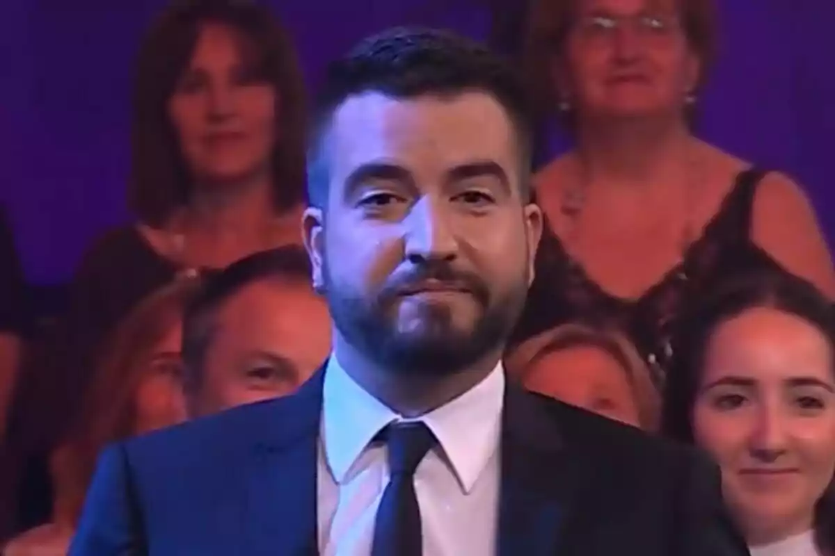 Ricard Ustrell amb barba i vestit fosc en primer pla amb públic desenfocat al fons en un ambient d’estudi de televisió de Col·lapse