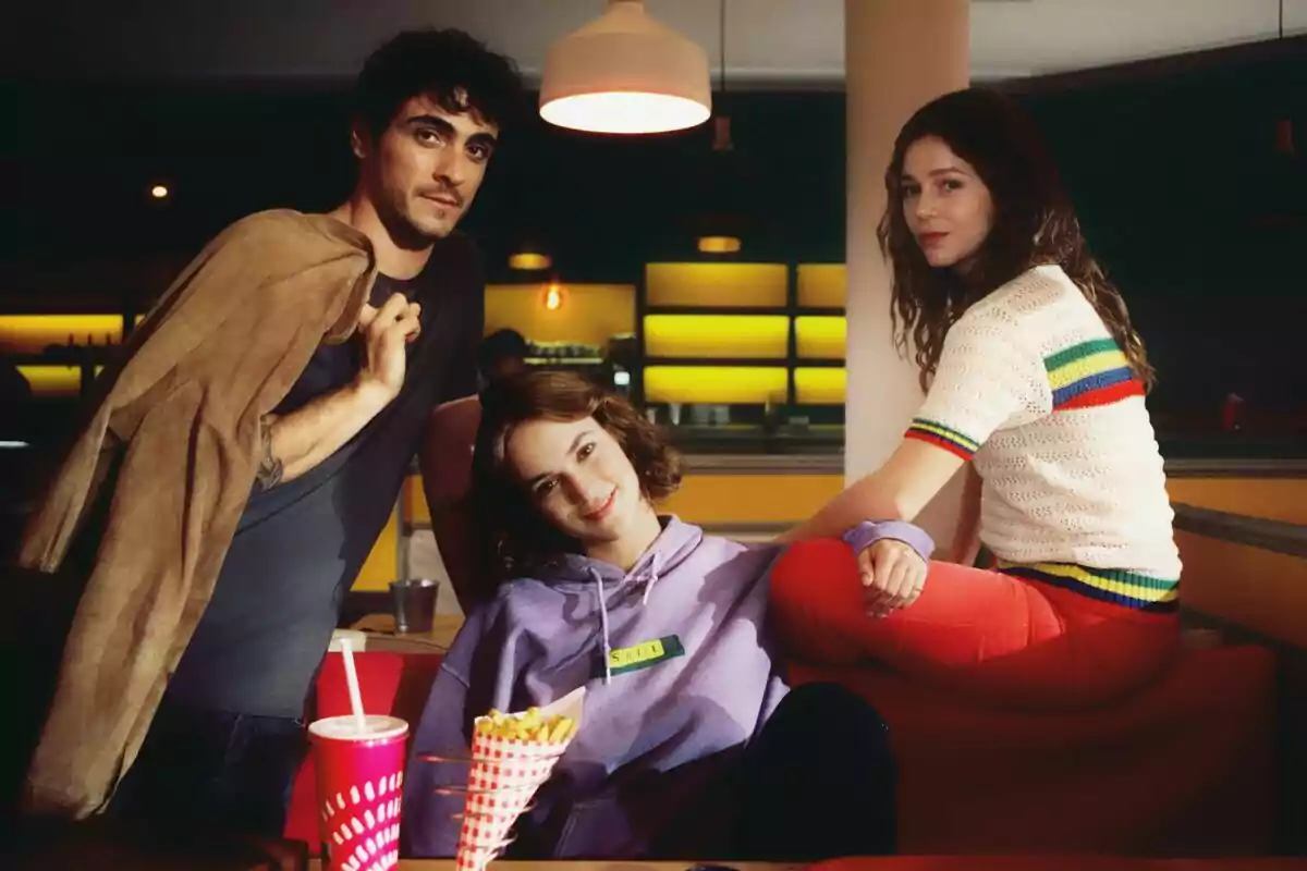 Alicia Falcó, Pablo Álvarez y Georgina Amorós jóvenes posan en el interior de una cafetería con ambiente retro, una de ellas está sentada en el respaldo del sofá, otra lleva una sudadera lila y la tercera sostiene una chaqueta sobre el hombro, en la mesa hay papas fritas y un vaso con popote en El mapa de los anhelos.