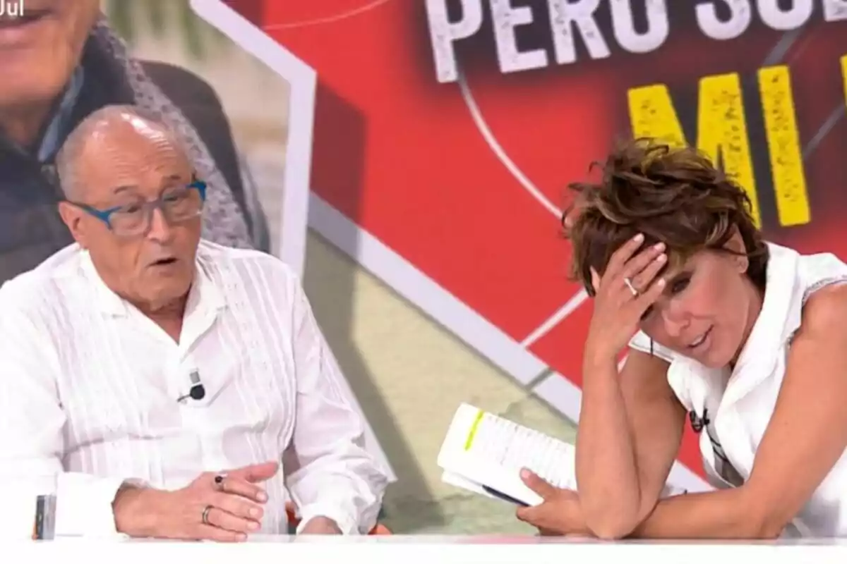 Dos personas sentadas en un set de Y ahora Sonsoles, una de ellas lleva gafas y habla mientras Sonsoles Ónega se cubre la frente con la mano y sostiene un cuaderno, ambos visten de blanco.