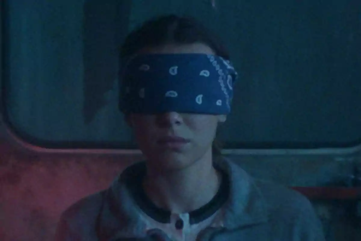 Millie Bobby Brown con los ojos cubiertos por una venda azul en un ambiente oscuro en Stranger Things.
