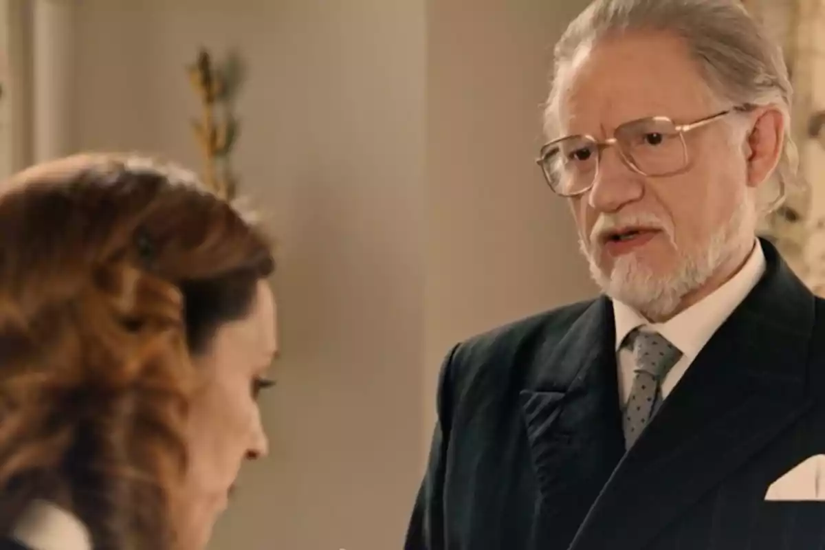 Damián con barba y gafas habla seriamente con Manuela de cabello castaño en un ambiente interior elegante. Captura de 'sueños de libertad' Damián con barba y gafas habla seriamente con Manuela de cabello castaño en un ambiente interior elegante. Captura de 'sueños de libertad'