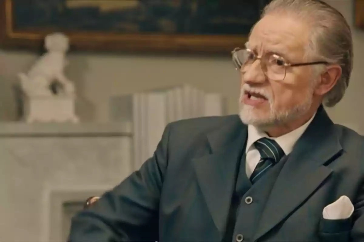 Damián amb ulleres i barba canosa vestit amb un vestit fosc i corbata de ratlles assegut en una habitació elegant. Captura de 'somnis de llibertat' Damián amb ulleres i barba canosa vestit amb un vestit fosc i corbata de ratlles assegut en una habitació elegant. Captura de 'somnis de llibertat'