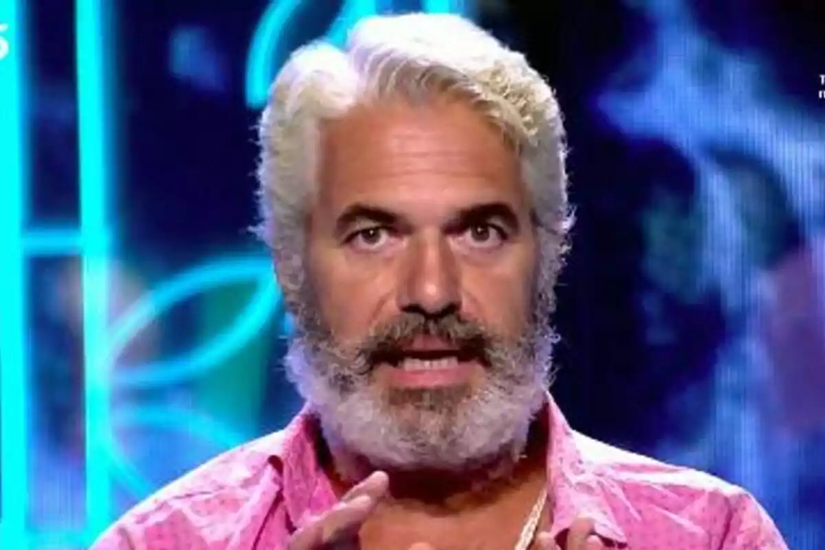 Agustín Bravo de cabell i barba canosos amb camisa rosa parlant davant d’un fons blau il·luminat a Supervivientes
