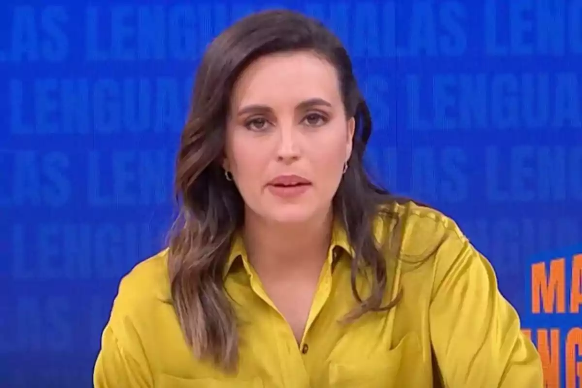 Aida Bao de cabello castaño y ondulado usando una camisa amarilla sentada frente a un fondo azul con letras desenfocadas en Malas Lenguas.