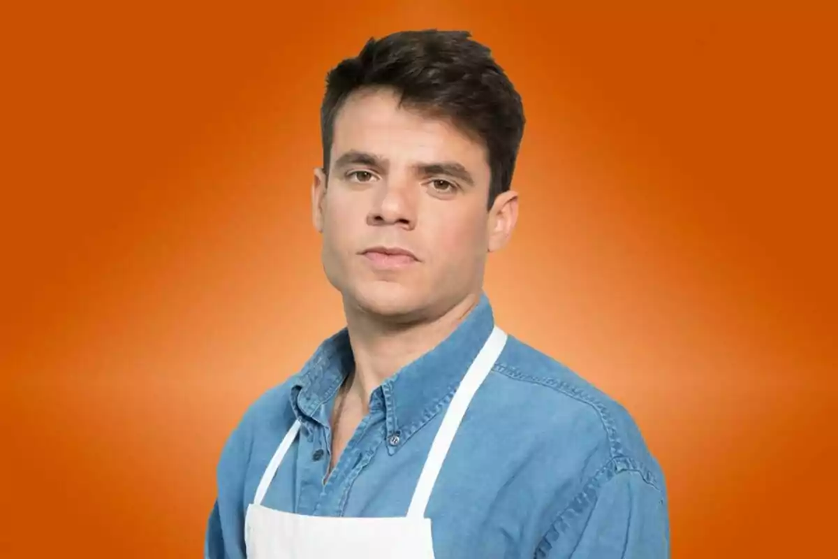 Alberto Gras joven con cabello oscuro, camisa de mezclilla azul y delantal blanco posando frente a un fondo naranja de MasterChef Alberto Gras joven con cabello oscuro, camisa de mezclilla azul y delantal blanco posando frente a un fondo naranja de MasterChef