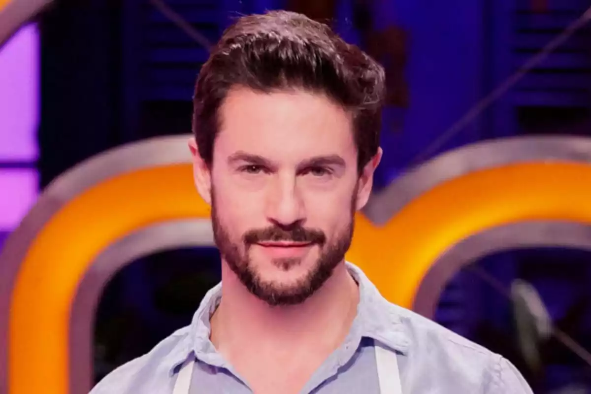 Alejo Sauras joven con barba y cabello oscuro, usando una camisa azul claro y un delantal blanco de MasterChef Celebrity, posando frente a un fondo colorido y desenfocado.