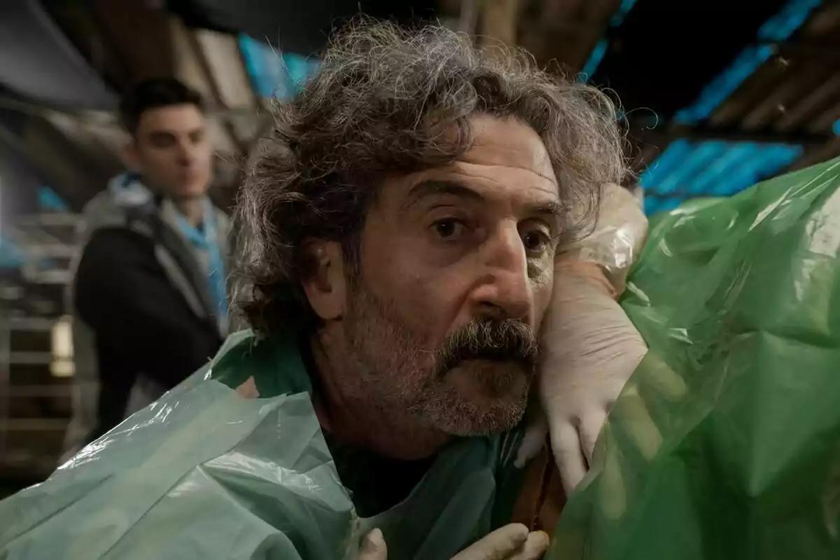 Luis Zahera de cabell arrissat i barba, amb bata i guants de làtex, observa atentament mentre realitza una tasca mèdica o científica en un entorn cobert amb plàstics verds a la sèrie Animal.