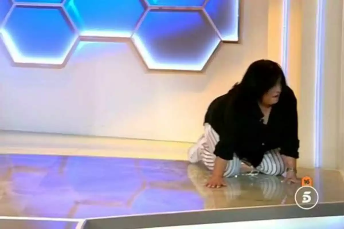 Mujer vestida con camisa negra y pantalón de rayas blancas y negras gateando sobre el suelo de un set de televisión con luces azules en el fondo en El diario de verano (El diario de Jorge) Mujer vestida con camisa negra y pantalón de rayas blancas y negras gateando sobre el suelo de un set de televisión con luces azules en el fondo en El diario de verano (El diario de Jorge)