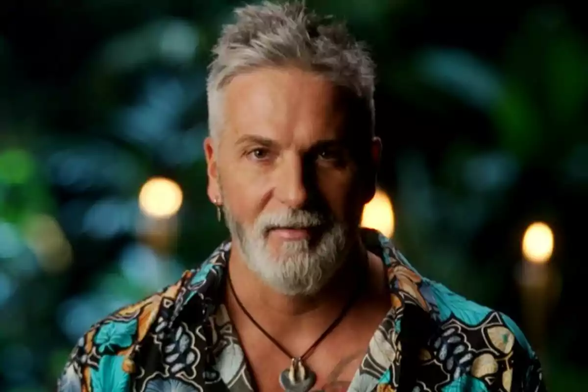 Carlos Alba de cabell i barba canosos amb camisa colorida i fons de llums desenfocades i vegetació a Supervivientes All Stars