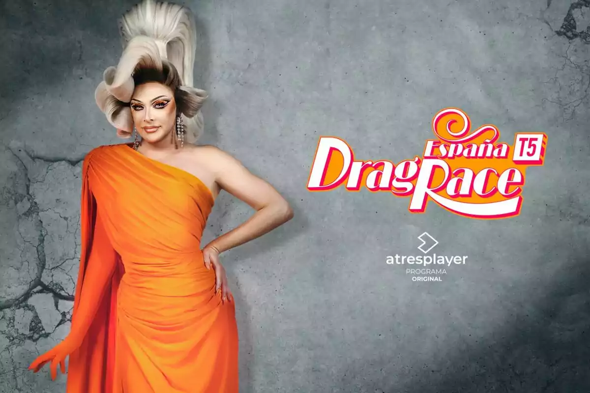 Supremme de Luxe con vestido naranja y peluca rubia voluminosa posando junto al logo de Drag Race España T5 sobre un fondo gris agrietado Supremme de Luxe con vestido naranja y peluca rubia voluminosa posando junto al logo de Drag Race España T5 sobre un fondo gris agrietado