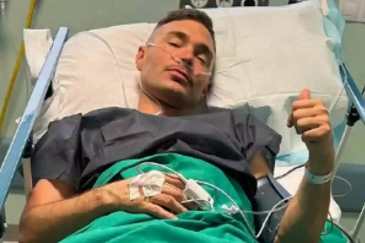 Cristian Suescun acostado en una cama de hospital con bata médica y oxígeno, levantando el pulgar en señal de ánimo