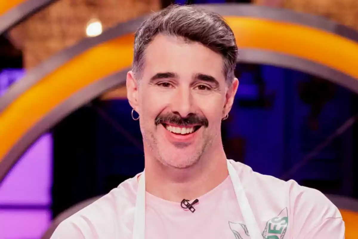 David Amor de cabell curt i canós amb bigoti i samarreta rosa somrient davant d’un fons colorit de MasterChef Celebrity.