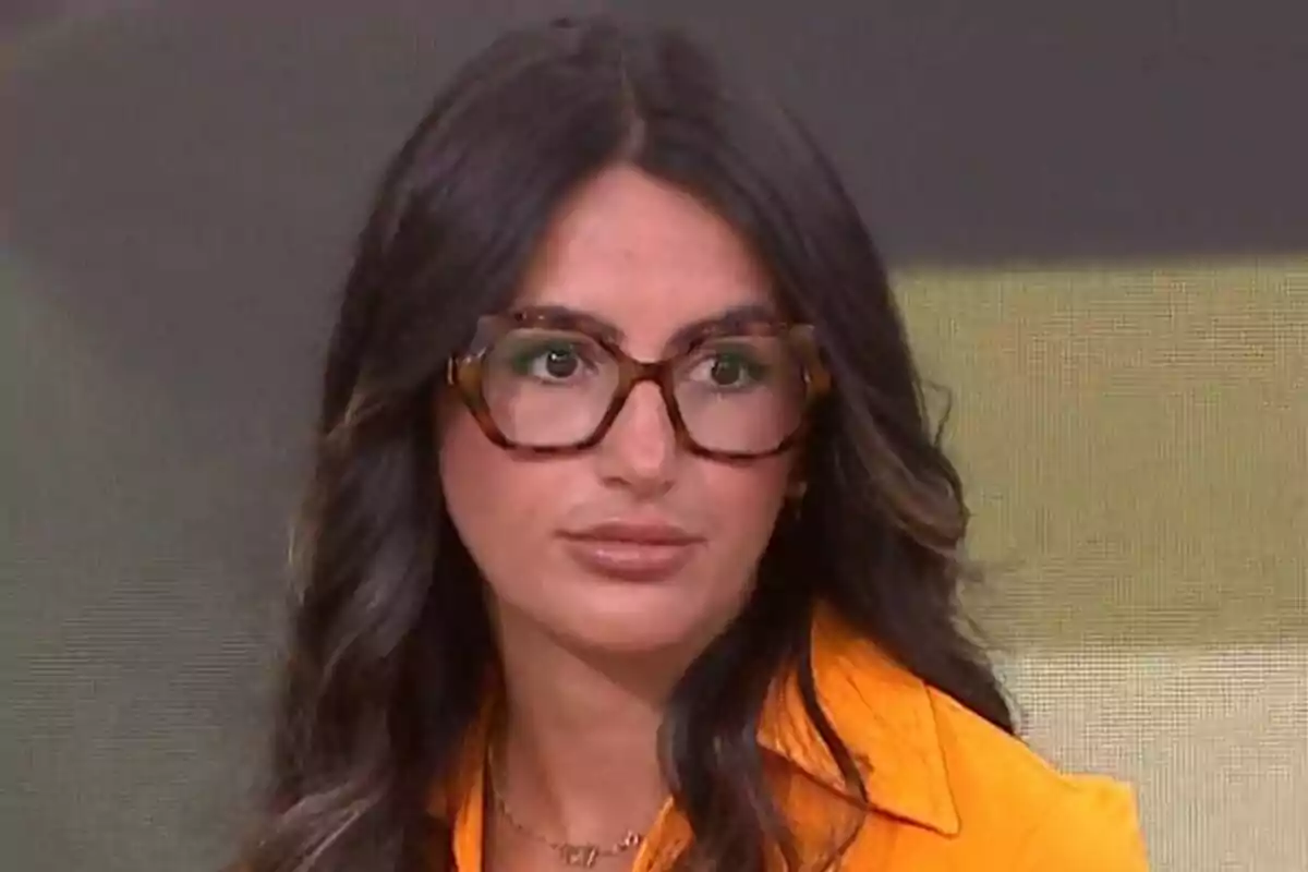Diana Higueras de cabell llarg i fosc amb ulleres grans i camisa taronja mirant cap a un costat a Espejo Público.