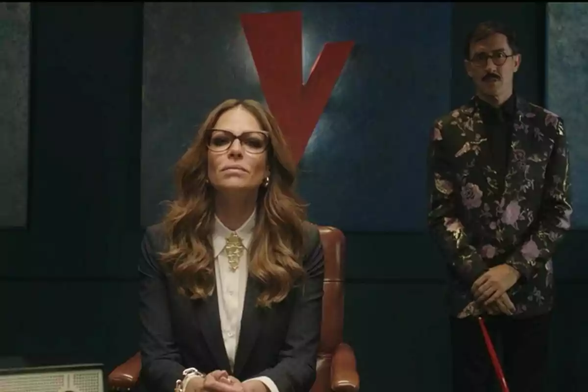 Eva González con gafas y cabello largo y ondulado está sentada en una silla de oficina mientras un hombre con bigote y saco floral está de pie detrás de ella frente a una pared azul con una gran letra V roja en el tráiler de La Voz. Eva González con gafas y cabello largo y ondulado está sentada en una silla de oficina mientras un hombre con bigote y saco floral está de pie detrás de ella frente a una pared azul con una gran letra V roja en el tráiler de La Voz.