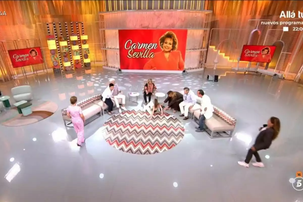 Un grup de persones amb la Terelu Campos al capdavant conversa en un plató de Fiesta i una persona està estirada a terra decorat amb tons taronges i daurats, amb una gran pantalla al fons que mostra la imatge d’una dona i el nom Carmen Sevilla. Un grup de persones amb la Terelu Campos al capdavant conversa en un plató de Fiesta i una persona està estirada a terra decorat amb tons taronges i daurats, amb una gran pantalla al fons que mostra la imatge d’una dona i el nom Carmen Sevilla.