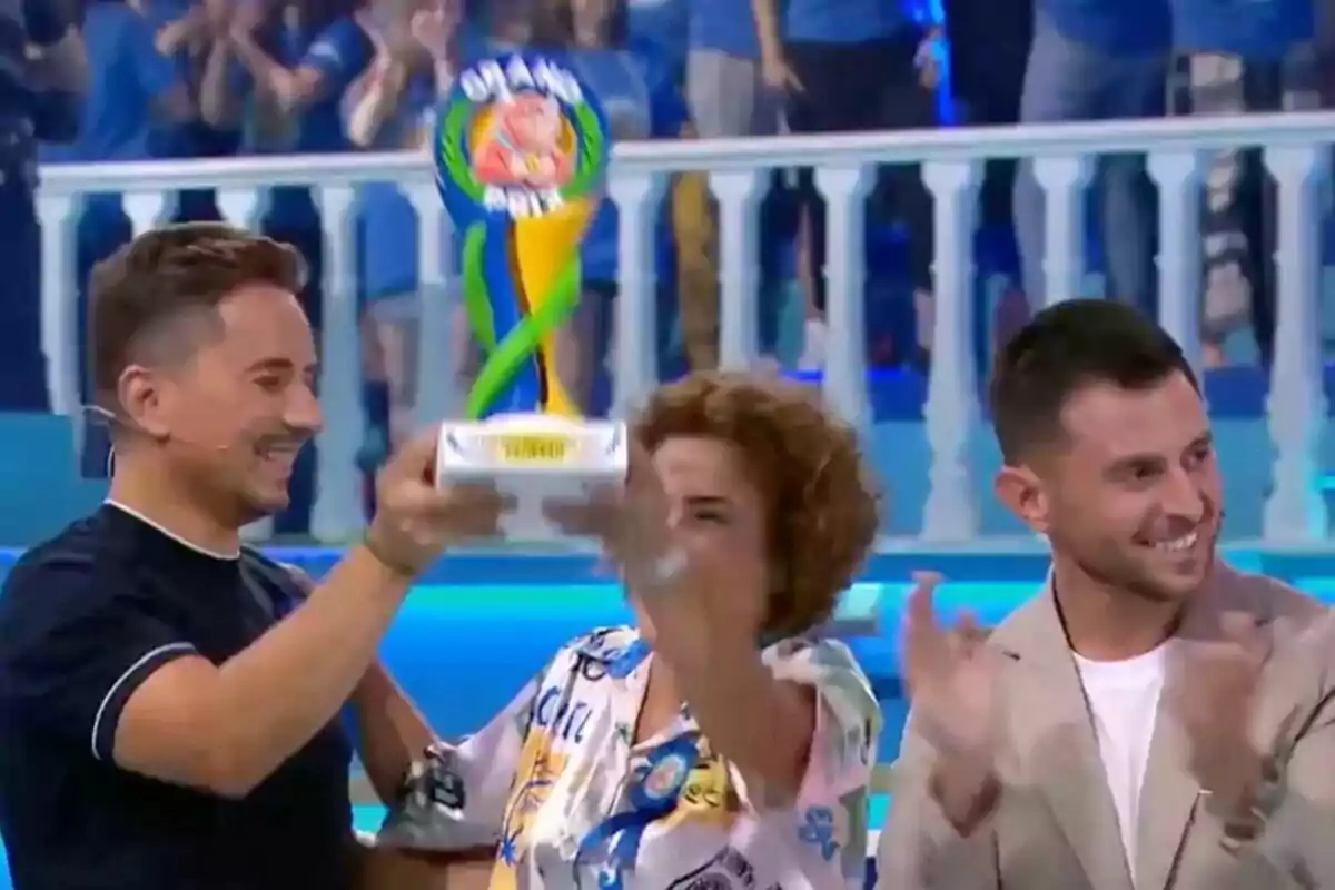 Tres personas sonríen, entre ellas Jorge Lorenzo, y celebran mientras una de ellas sostiene un trofeo colorido de El Grand Prix en un ambiente festivo con público al fondo Tres personas sonríen, entre ellas Jorge Lorenzo, y celebran mientras una de ellas sostiene un trofeo colorido de El Grand Prix en un ambiente festivo con público al fondo