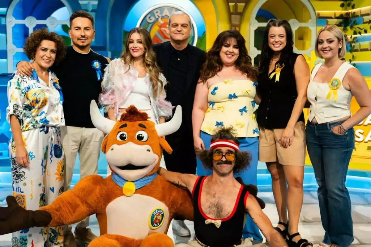 Un grupo de personas formado por Ramón García, Lalachus, Ángela Fernández, Wilbur, Jorge Lorenzo y Adriana Torrebejano sonríe posando junto a una botarga de toro y un hombre disfrazado con peluca y gafas en un colorido set de televisión en El Grand Prix.