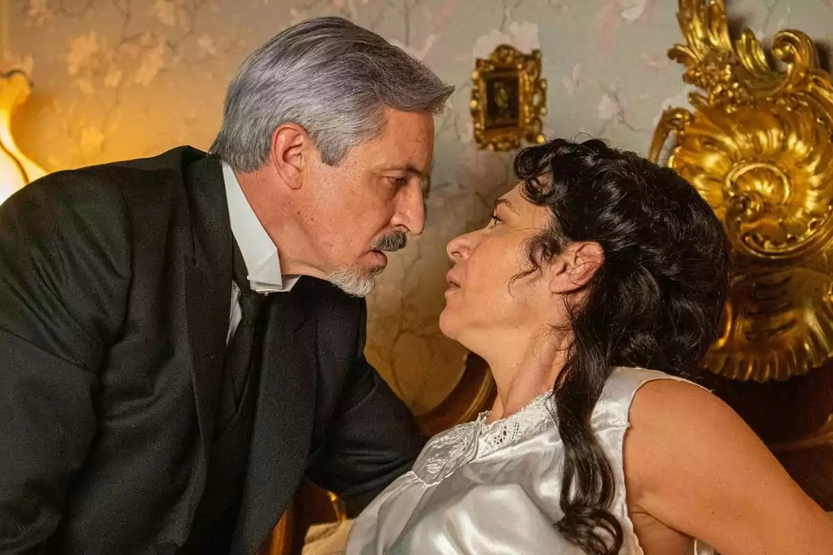 Fernando Coronado de cabello canoso y barba se inclina hacia Isabel Serrano de cabello oscuro que está sentada en una cama con cabecera dorada, ambos se miran intensamente en una habitación decorada con papel tapiz floral en La Promesa. Fernando Coronado de cabello canoso y barba se inclina hacia Isabel Serrano de cabello oscuro que está sentada en una cama con cabecera dorada, ambos se miran intensamente en una habitación decorada con papel tapiz floral en La Promesa.