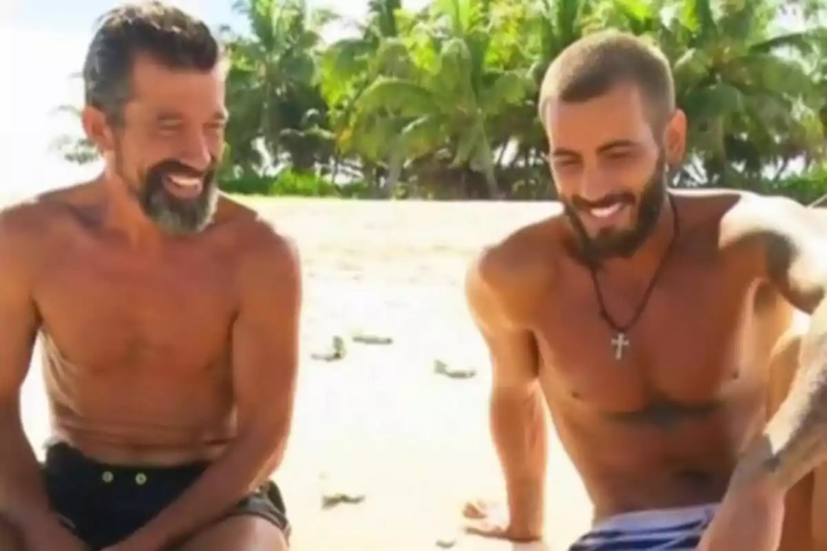 Iván González i José Luis Losa somrients i sense samarreta asseguts a la sorra d’una platja amb palmeres al fons a Supervivientes