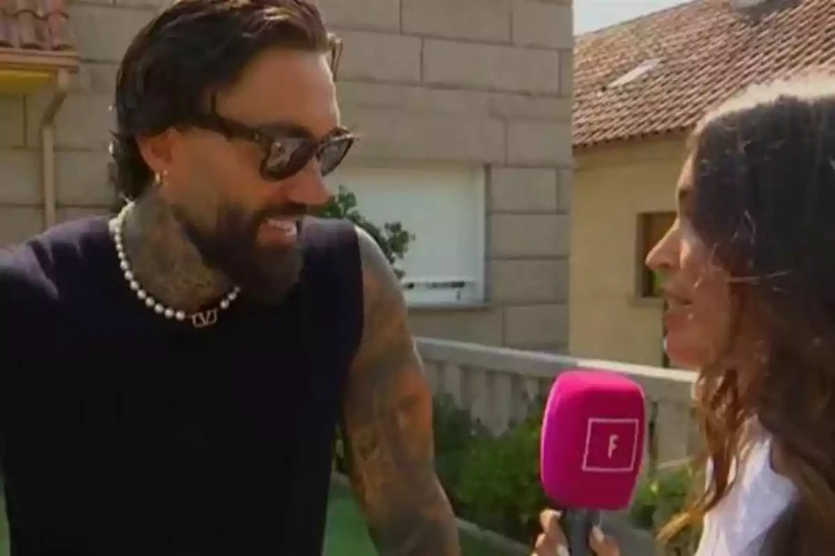 Juan Faro con gafas de sol y tatuajes sonríe mientras es entrevistado por una reportera que sostiene un micrófono rosa con una letra F de Fiesta en el exterior de una casa. Juan Faro con gafas de sol y tatuajes sonríe mientras es entrevistado por una reportera que sostiene un micrófono rosa con una letra F de Fiesta en el exterior de una casa.